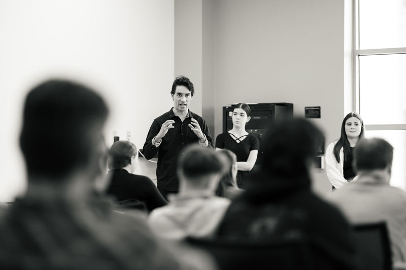 A person stands and speaks to a seated group about management; two others sit nearby.