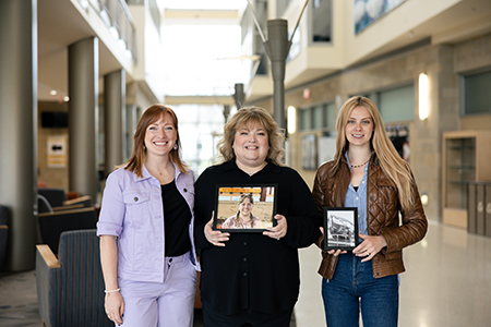 Three women stand indoors, smiling at the camera. The woman in the center holds a framed photo of a young woman, and the woman on the right holds a small framed black-and-white photo of a building.