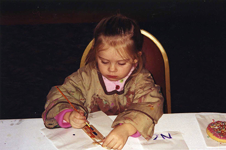 A young child wearing a paint-splattered smock sits at a table, focused on painting with a brush. There are papers and a decorated cookie on the table in front of her.