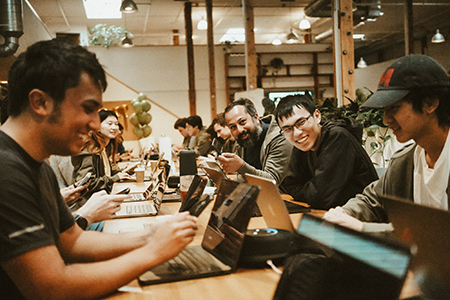 A group of people sit together at a long wooden table, working on laptops and smiling in a bright, modern, open office space with plants and shelves in the background.