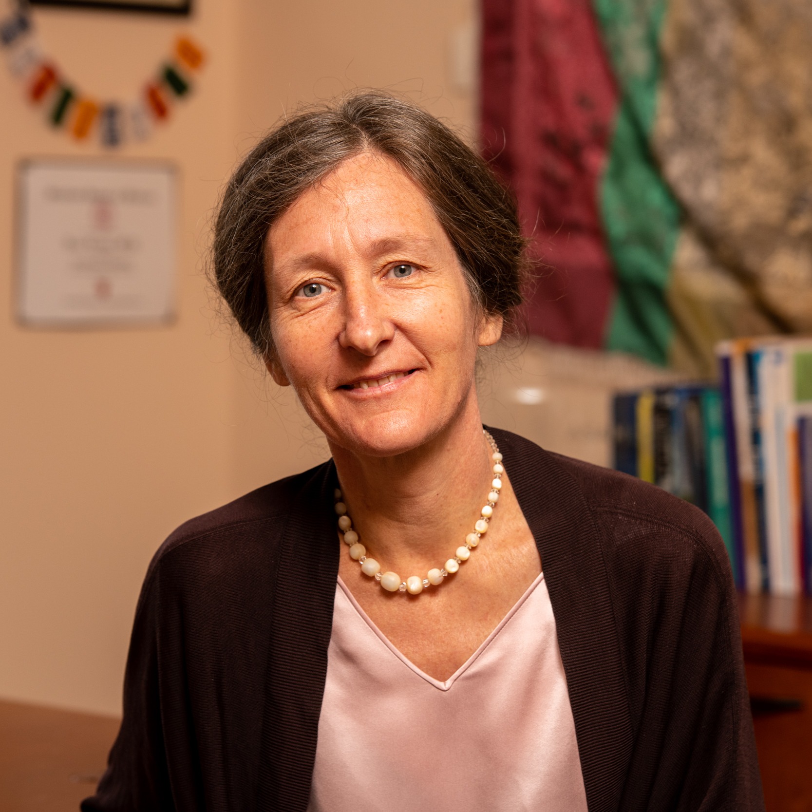 A middle-aged woman with brown hair tied back, wearing a pearl necklace, light pink blouse, and dark cardigan, smiles while sitting indoors, surrounded by books and a framed certificate that hint at her past events and activities.