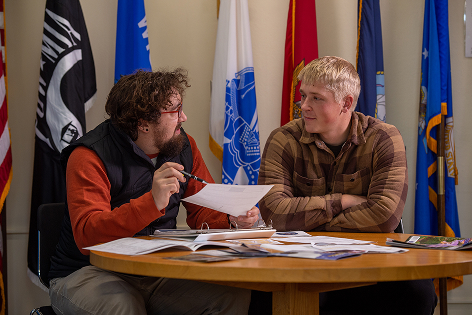 Two men sit at a table discussing academic microcredentials paperwork in front of several flags.