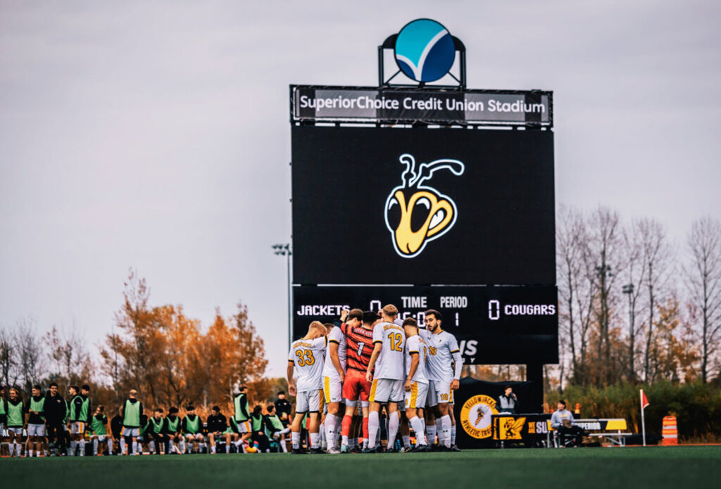 A soccer team in yellow and white uniforms huddles on the field in front of a large scoreboard at SuperiorChoice Credit Union Stadium, with players and trees visible in the background.