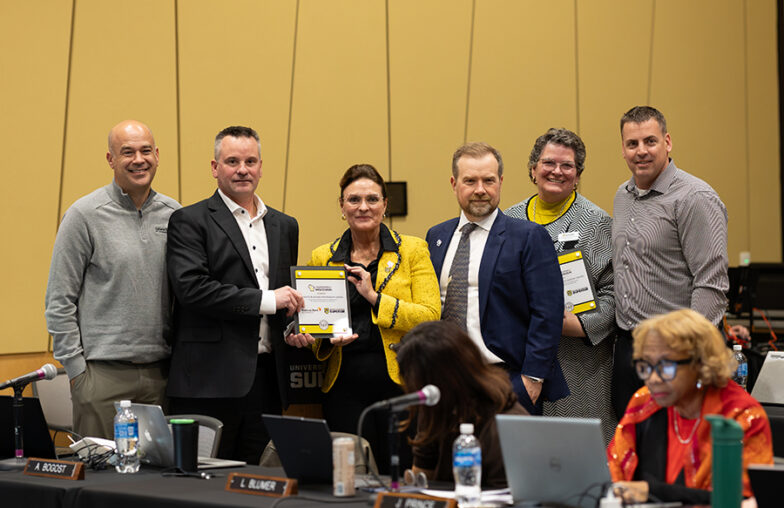 A group of people standing behind a table, one woman holding an award certificate.