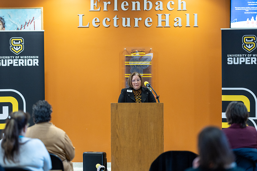 A woman speaks at a podium in Erlenbach Lecture Hall at the University of Wisconsin-Superior, with attendees seated and two university banners displayed on either side.