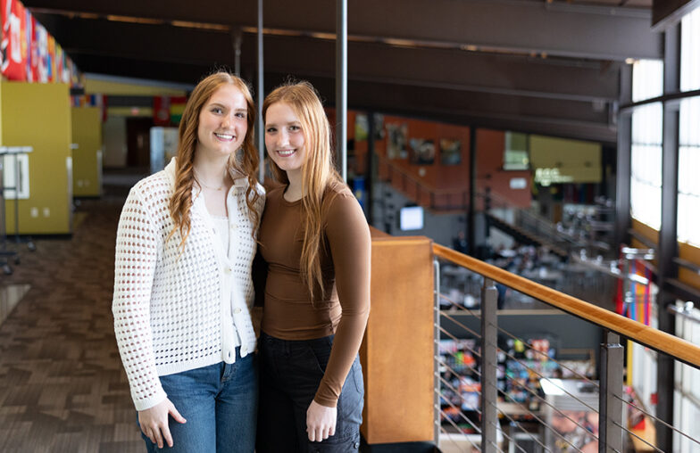 Two young women stand side by side in a modern building with large windows and flags.