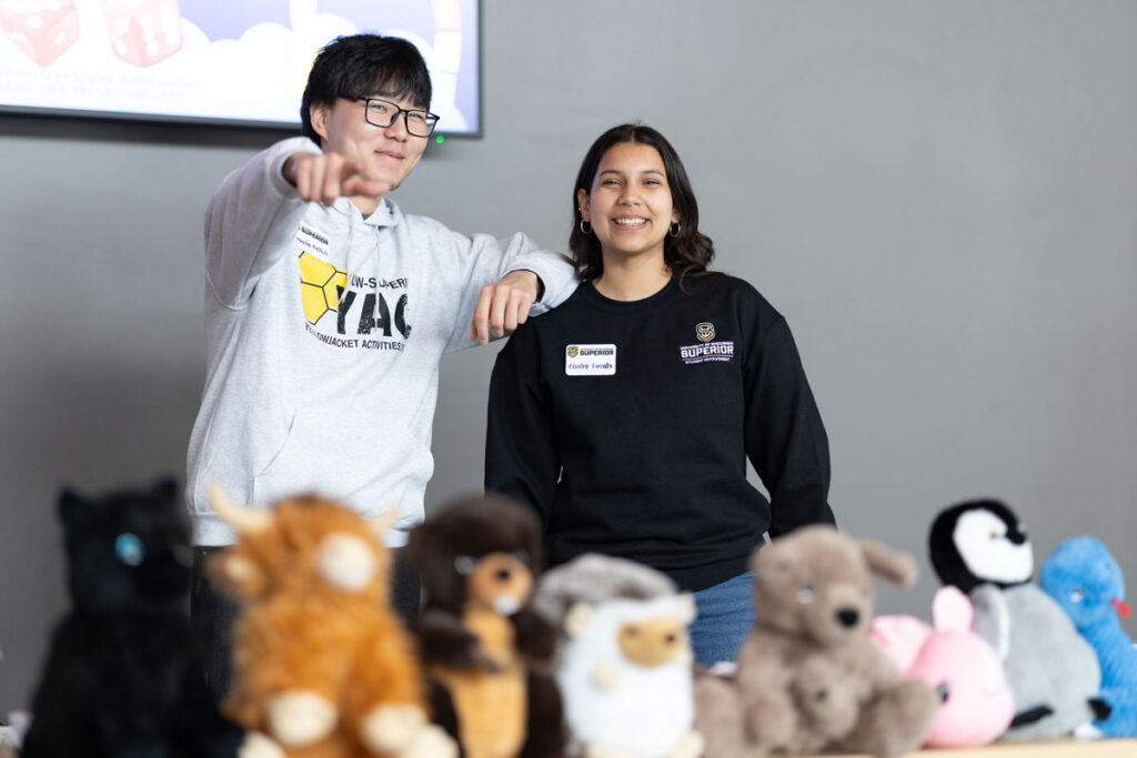 Two people stand behind a table displaying various plush toys, representing student employment opportunities. One person points towards the camera, both are smiling.