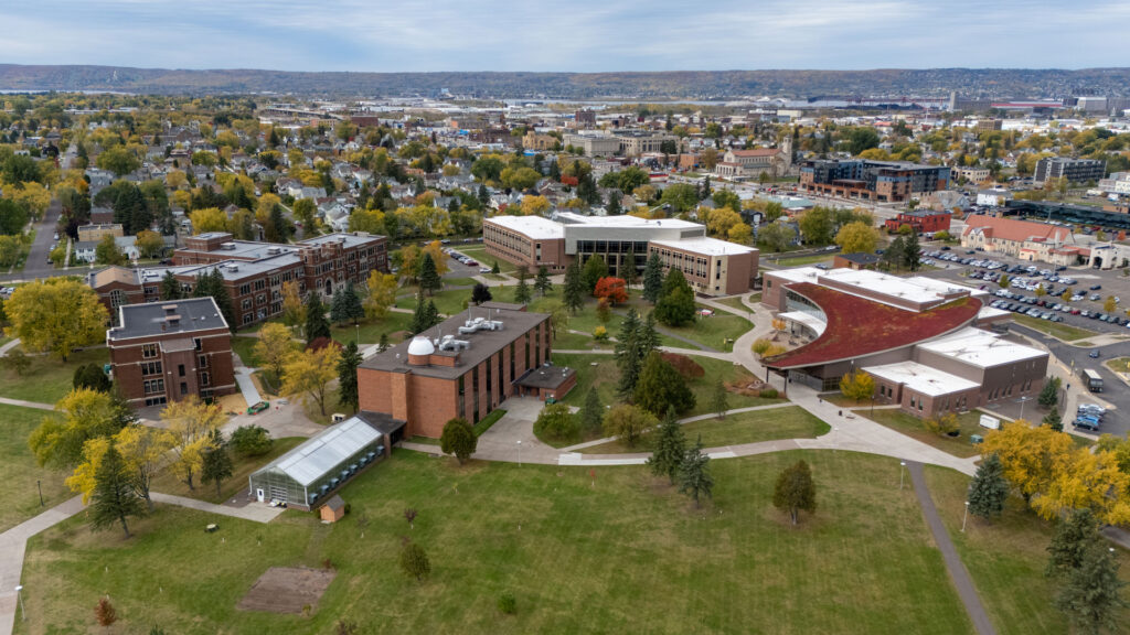 Aerial view of the University of Wisconsin-Superior campus with brick and modern buildings, pathways, autumn foliage, and green spaces; the cityscape and hills form a scenic backdrop under a cloudy sky. Learn more: About UW-Superior.