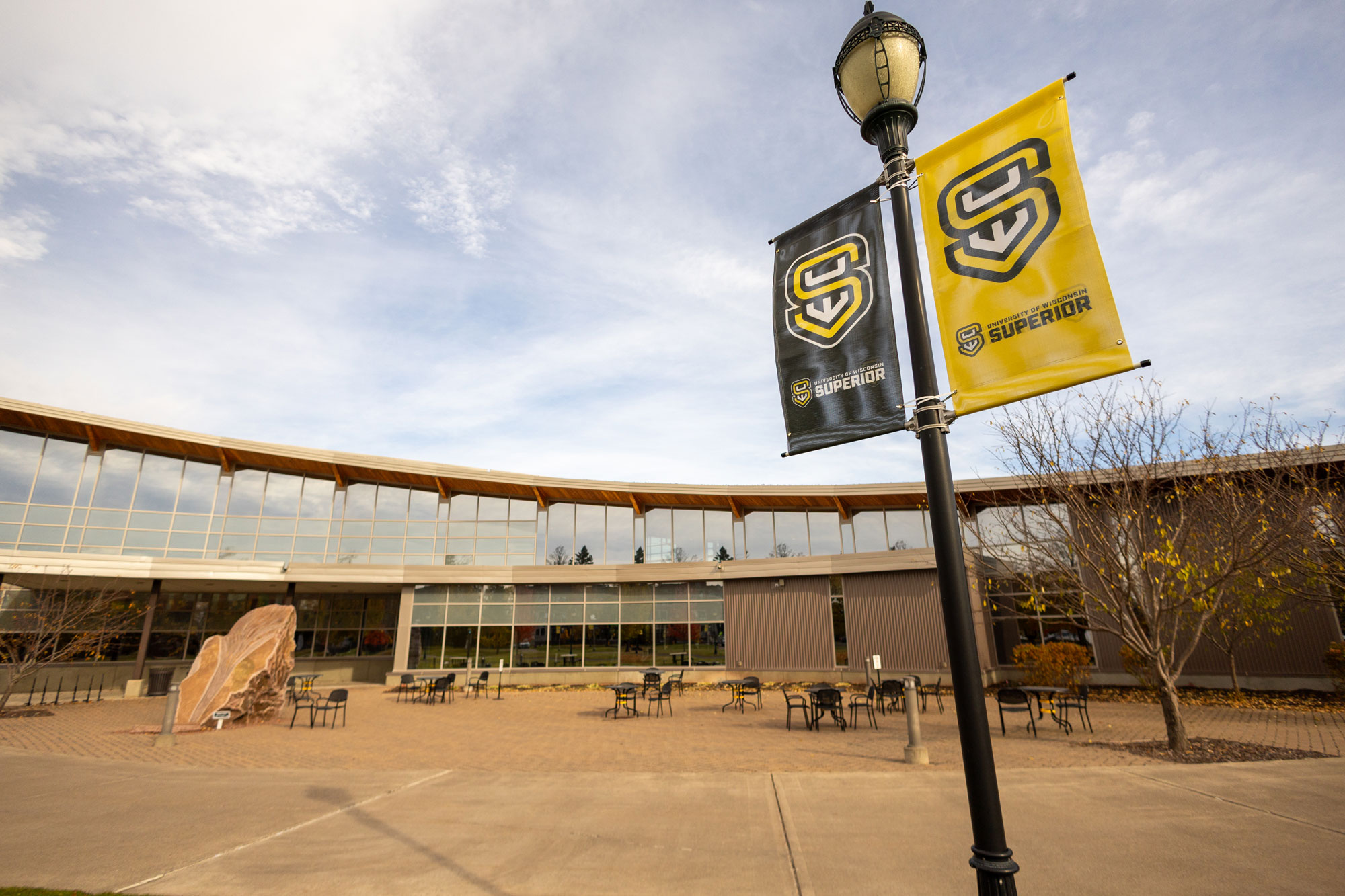 Outdoor view of a modern building at the University of Wisconsin-Superior with large windows, patio tables and chairs, a rock sculpture, and two banners displaying the Superior logo. Leafless trees add to the inviting student atmosphere.
