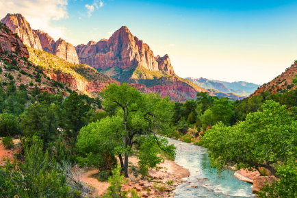 A scenic view of a river winding through lush green trees, with tall red rock mountains in the background under a blue sky at sunset.
