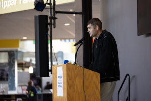 Man speaking at a podium with a microphone indoors; sign reads campus Superior Pathway..