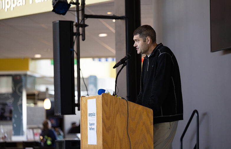 Man speaking at a podium with a microphone indoors; sign reads campus Superior Pathway..