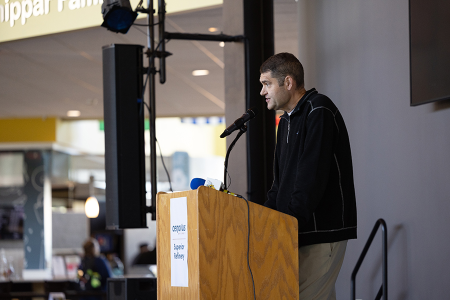 Man speaking at a podium with a microphone indoors; sign reads campus Superior Pathway..