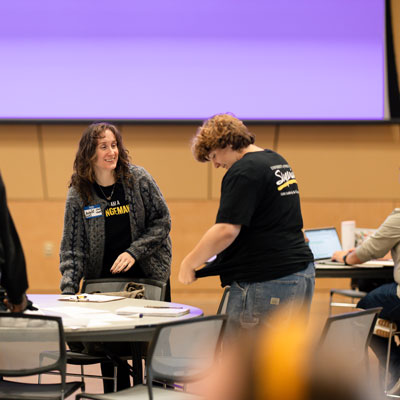 Two people chat near a round table in a conference room, perhaps discussing ideas for Youth Camps. One wears a name tag and gray sweater, the other a black t-shirt. A laptop, chairs, and a large blank screen are visible in the background.