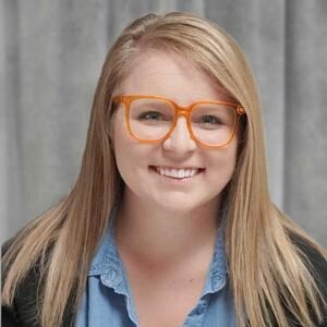 A woman with straight blonde hair, wearing orange glasses, a blue button-up shirt, and a black cardigan, smiles in front of a gray curtain background.