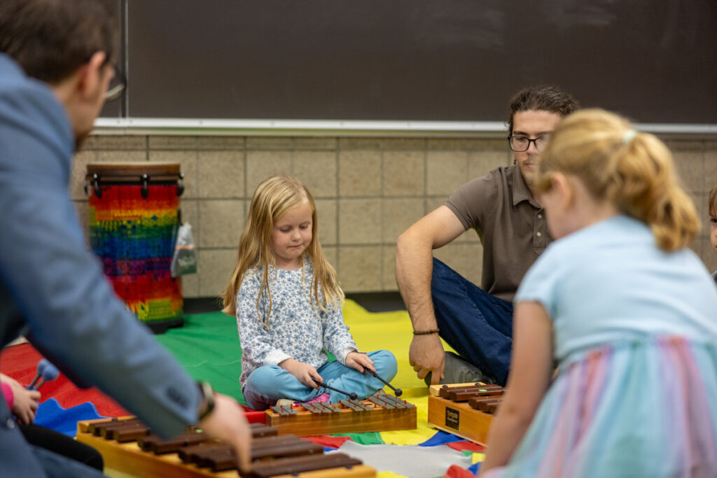 Children and adults sit in a circle on a colorful mat, playing xylophones together. A young girl is focused on her instrument, while others watch and participate in the music activity.