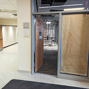 A hallway with an open door reveals a room under construction, with exposed metal framing and a ladder inside. Brown paper covers the adjacent window. The hallway floor is tiled and brightly lit.