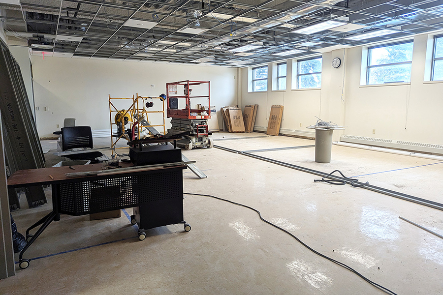 A large room under renovation with exposed ceiling tiles, construction equipment, scattered furniture, building materials, and a few windows letting in natural light.