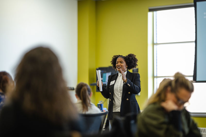 A woman in a blazer stands and speaks, holding papers in one hand, in front of a group discussing UW-Superior graduate programs in a brightly lit room with green walls and large windows.