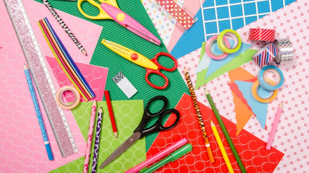 Colorful craft supplies on a table, including patterned paper, scissors, pencils, pens, washi tape, and striped straws, all arranged in a scattered, creative layout.