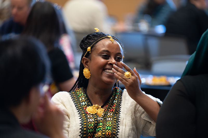 A woman dressed in traditional attire and gold jewelry smiles joyfully, covering part of her face with her hand. She sits indoors among a group, reflecting the spirit of multicultural initiatives at a lively social or cultural event.