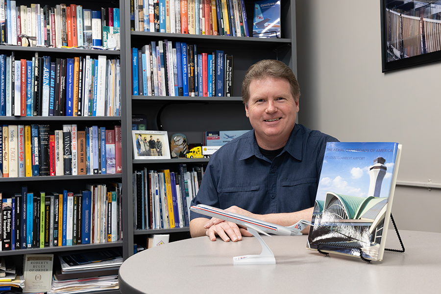 Man sitting at a desk with a model airplane, book, and bookshelves in the background.
