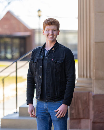 Young man with red hair stands outside near a stone building, smiling at the camera.