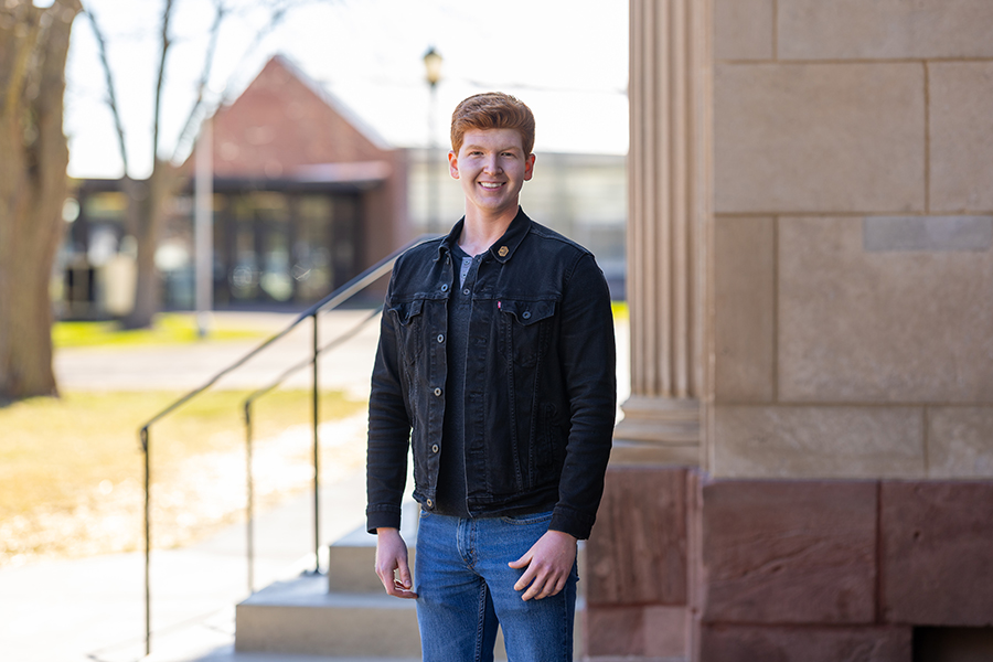 Young man with red hair stands outside near a stone building, smiling at the camera.