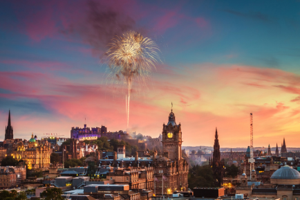 Fireworks light up the sky over Edinburgh, Scotland at sunset, with historic buildings, including the Balmoral Hotel clock tower and Edinburgh Castle, visible in the cityscape.