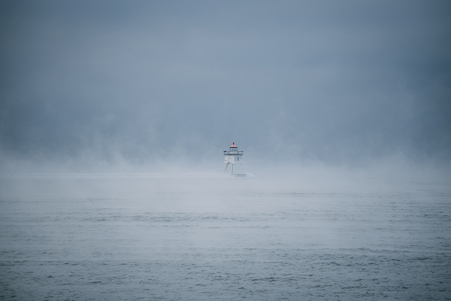 A white lighthouse with a red light stands on a pier, partially shrouded in mist, overlooking a calm, fog-covered body of water under an overcast sky.