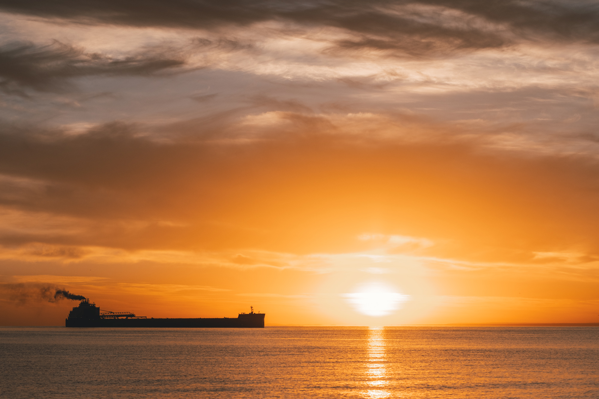 A large cargo ship sails across a calm sea at sunset, emitting smoke, with the sun low on the horizon and orange clouds filling the sky, adding an air of tranquility to just about everything in view.