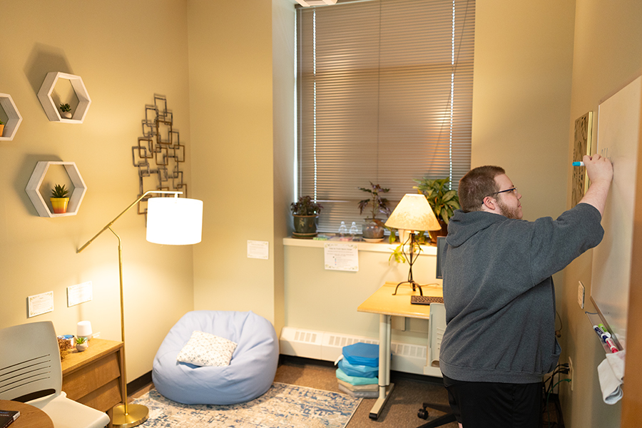 Person writing on a whiteboard in a cozy office with plants, lamp, and a bean bag chair.