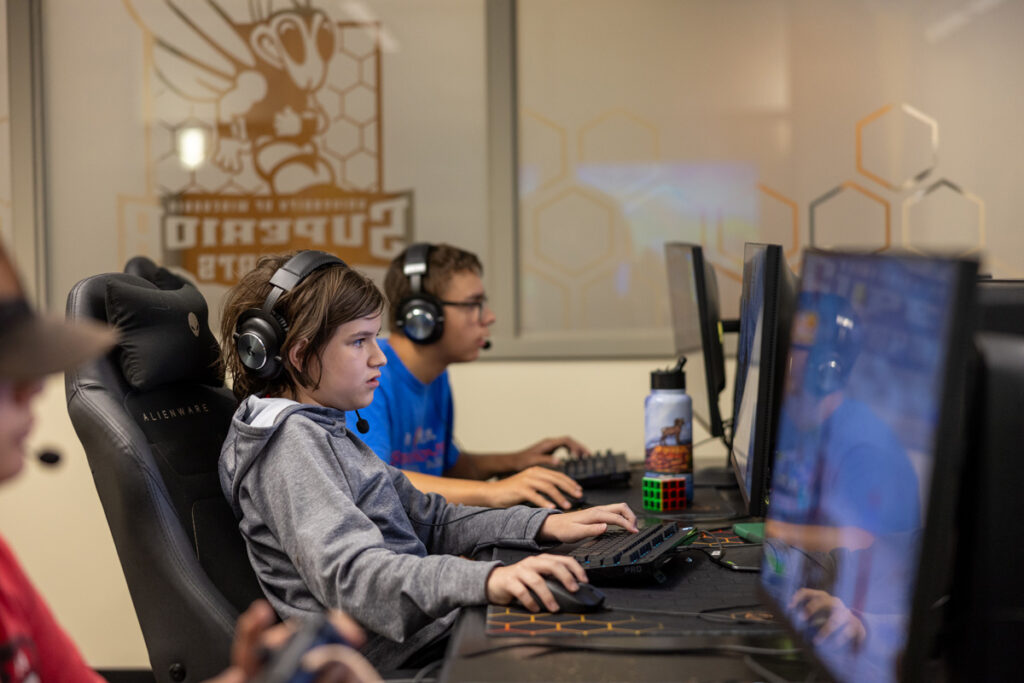 Three young people wearing headsets sit at desks, focused on computer screens while playing a video game at a vibrant summer camp, surrounded by a large Superio Esports logo and honeycomb patterns.