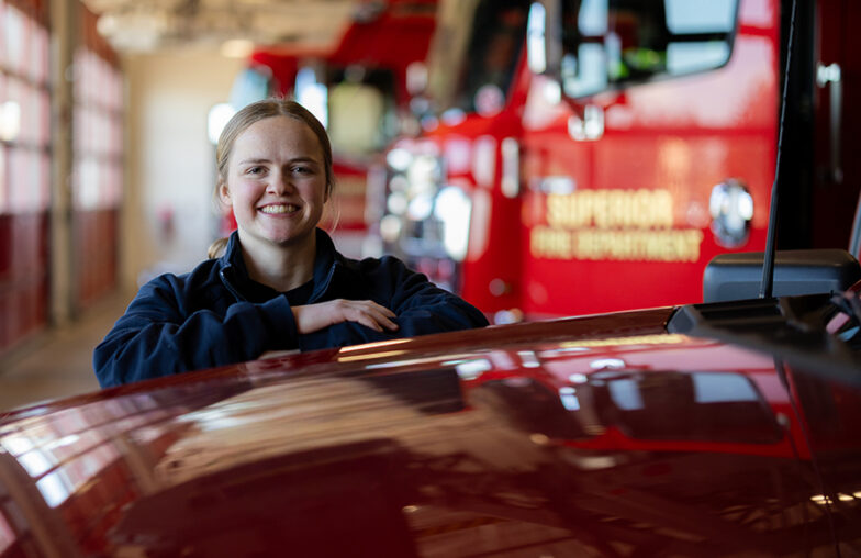 A firefighter stands and smiles in a fire station next to a red fire truck.