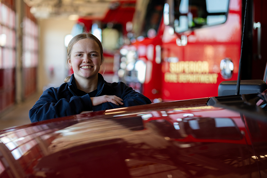 A firefighter stands and smiles in a fire station next to a red fire truck.