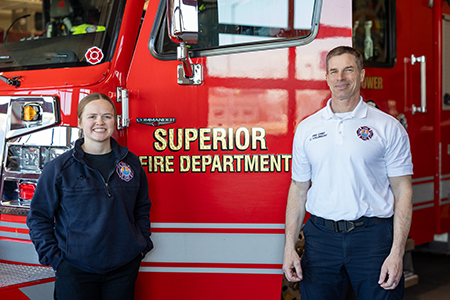Two firefighters stand in front of a red Superior Fire Department fire truck in a garage.