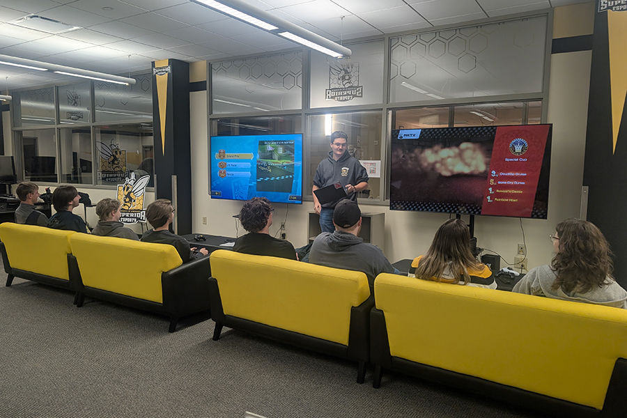 A person stands in front of two gaming screens as a group sits on yellow sofas.