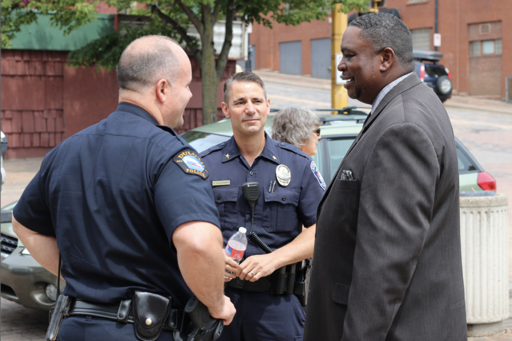 Two police officers in uniform stand outside talking to a man in a dark suit. One officer holds a water bottle. They appear to be having a friendly conversation near a parked car on a city street.
