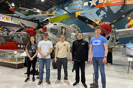 Five people stand in front of vintage airplanes in an aviation museum exhibit.