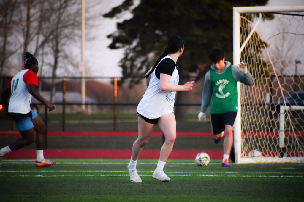 Three people play soccer on a field. One player in green, acting as goalie, kicks the ball while another in a white jersey runs toward the goal, and a third player in red shorts approaches from the left.
