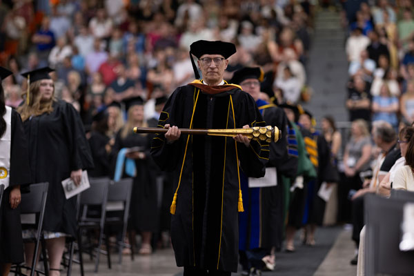 Faculty-Staff-Giving-web A person in academic regalia holds a ceremonial mace at a graduation ceremony indoors.