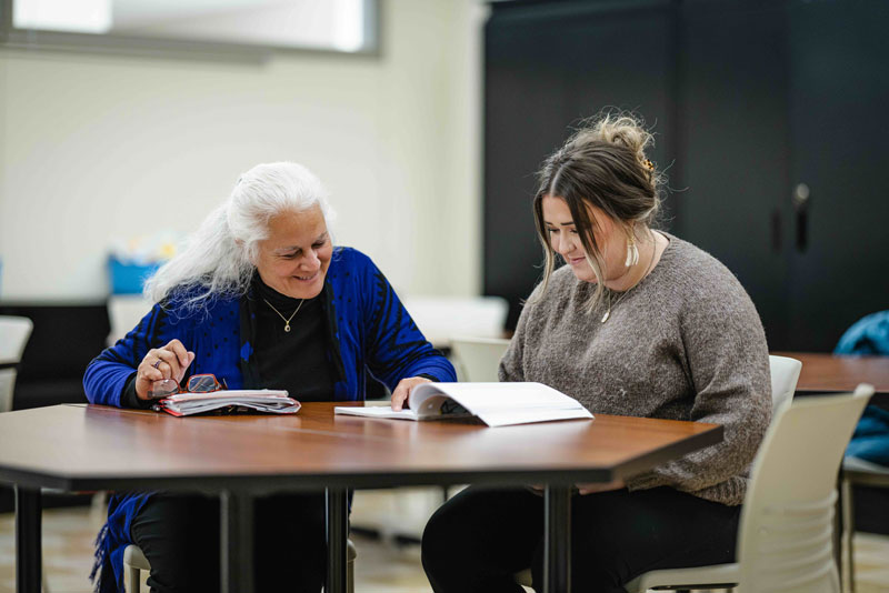 Two women sit at a table, smiling and looking at an open book together in a bright room, suggesting a study or mentoring session related to graduate programs.