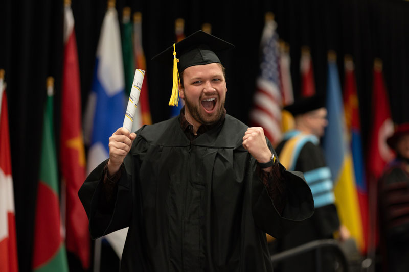 A joyful graduate in a cap and gown celebrates academic success while holding a diploma, with international flags and other graduates visible in the background.