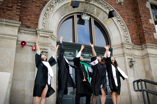 Five graduates in caps and gowns celebrate graduation by tossing hats before a brick school.