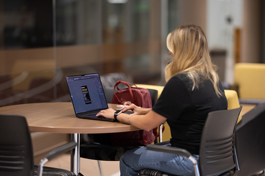 Person typing on a laptop at a round table with a red bag and empty chairs nearby.