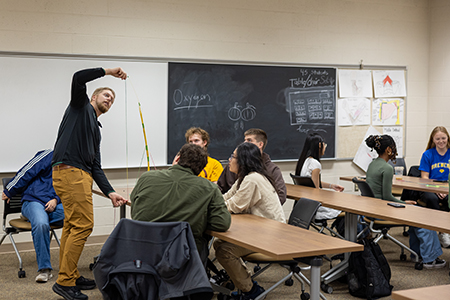A teacher demonstrates a science experiment with a string and weight to a group of attentive students seated at tables in a classroom with a chalkboard and posters on the wall.