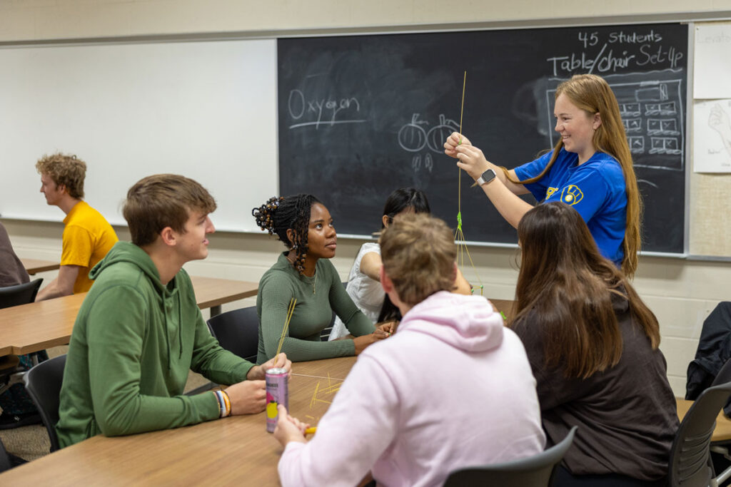 A group of students sits at desks in a classroom, watching as a standing student in a blue shirt demonstrates an activity with sticks—an example of inclusive teaching methods. A chalkboard with drawings and words is in the background.