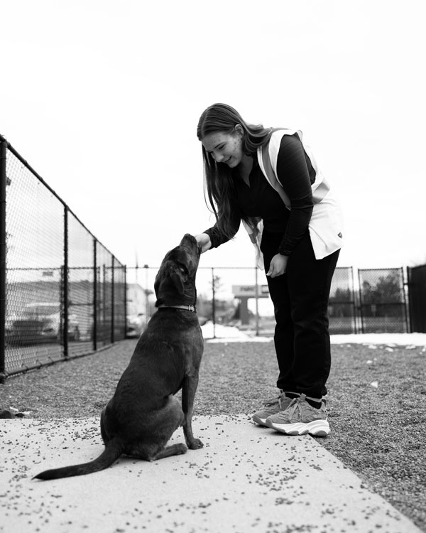 A woman practices psychology as she pets a dog sitting near a fenced outdoor area on the sidewalk.