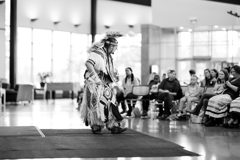 A person in traditional regalia dances indoors as an indigenous studies audience observes.