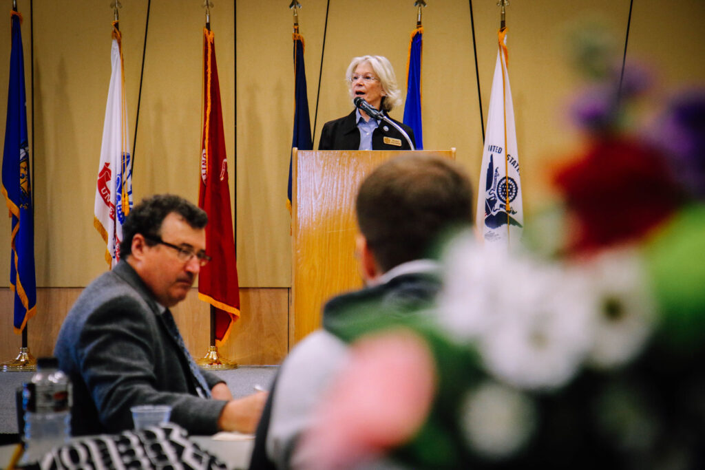 A woman speaks at a podium during annual events, framed by several flags, while two men sit at a table in the foreground. The focus is on the speaker, with the audience slightly blurred.